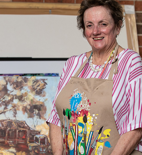 Cheryl wearing a painters apron stands in front if colourful painted canvases.
