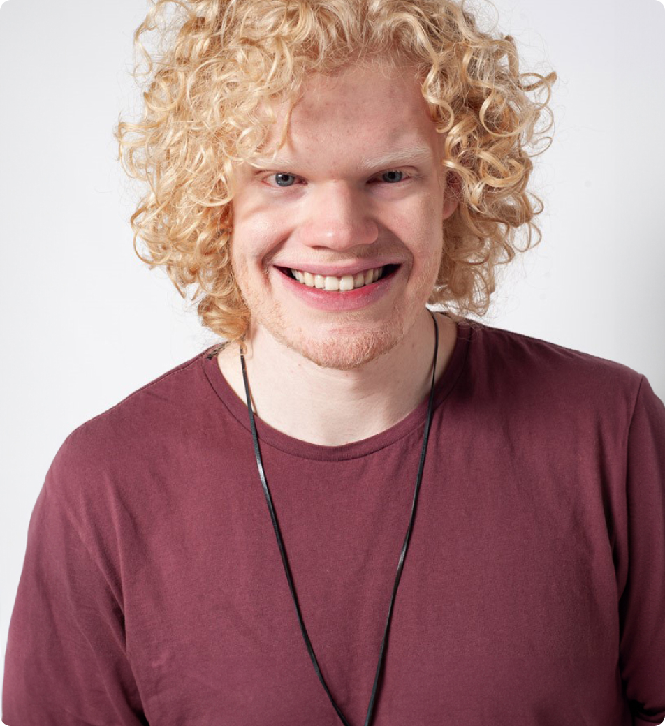 Image features a young man with curly blond hair and a wide smile. He's wearing a maroon T-shirt and a thin black necklace. The background is plain and light-coloured.