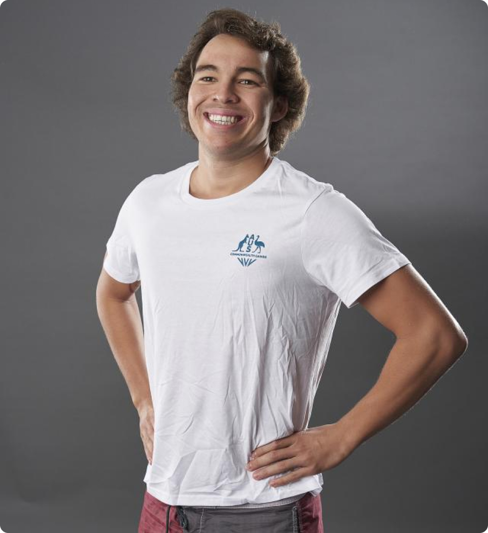 Image features a young man standing against a grey background. He has a friendly expression, with a broad smile and slightly raised eyebrows, giving a relaxed and cheerful vibe. He's wearing an AUS Commonwealth Games white t-shirt and board shorts.