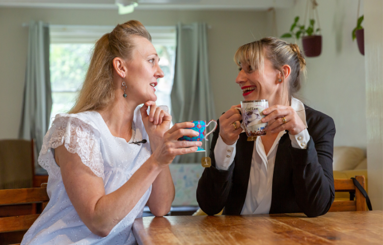 Two women sit at a dining table holding cups of tea and smiling in conversation with each other. The woman on the left has lived experience with vision loss.