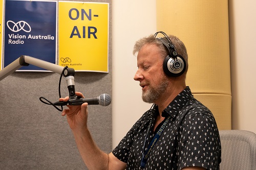 A man sits in a radio studio with a microphone in front of his face and an on air sign above his head