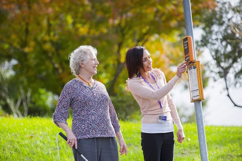 An older female Vision Australia client stands holding a white can as a Vision Australia staff member directs her to a bus stop