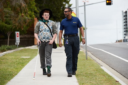 A Vision Australia client using a whiote can walks along a footpath with an orientatio and mobility specialist