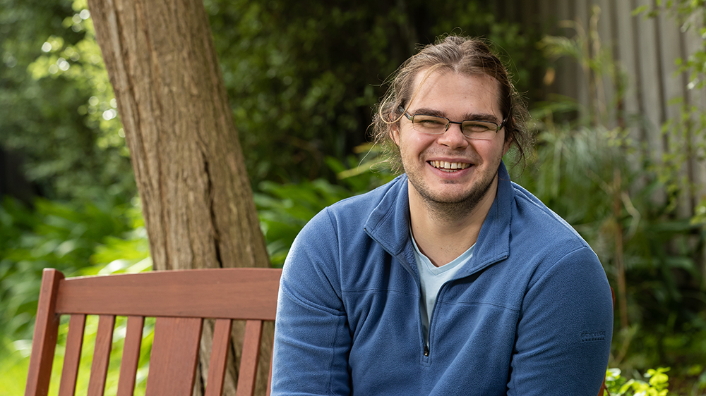 Ben is smiling to camera, seated on a park bench