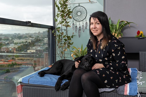 A woman sits on a balcony with a black labrador dog guide resting on her lap