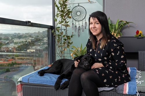 A woman sits on a balcony with a black labrador dog guide resting on her lap
