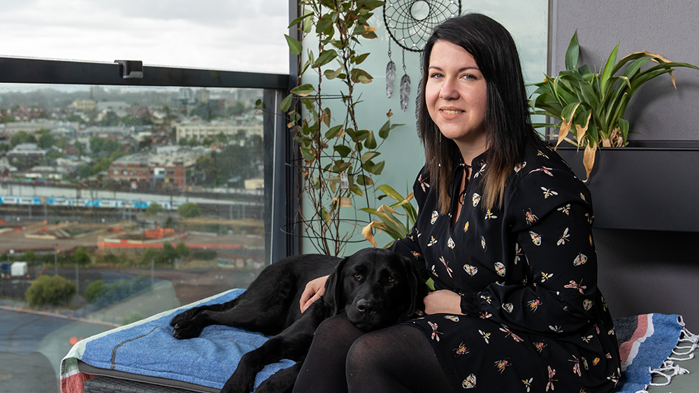 Brittee is smiling and sitting beside her black Labrador dog, on a balcony with urban views