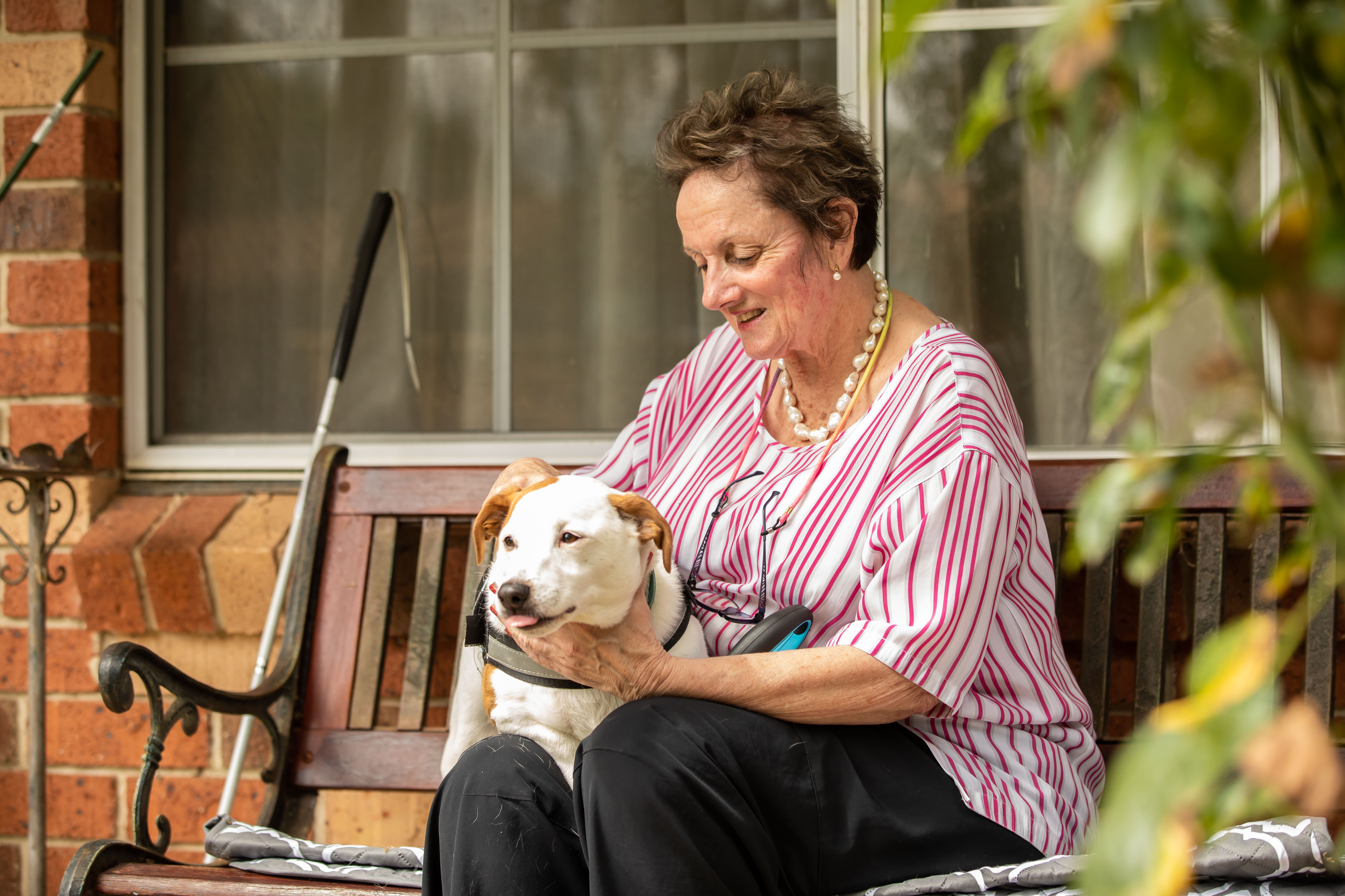 Vision Australia client Cheryl is smiling as she sits on a bench in her front garden with her dog guide.