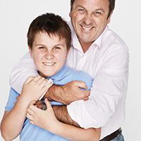 Father playfully hugs his son, they are smiling at camera.