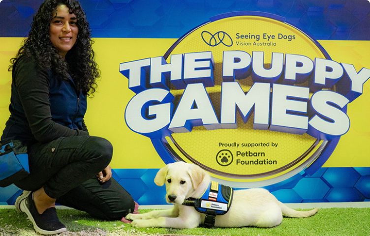 Seeing eye dog puppy Isobel lies at the feet of her trainer in front of big 'The Puppy Games' sign ready to compete