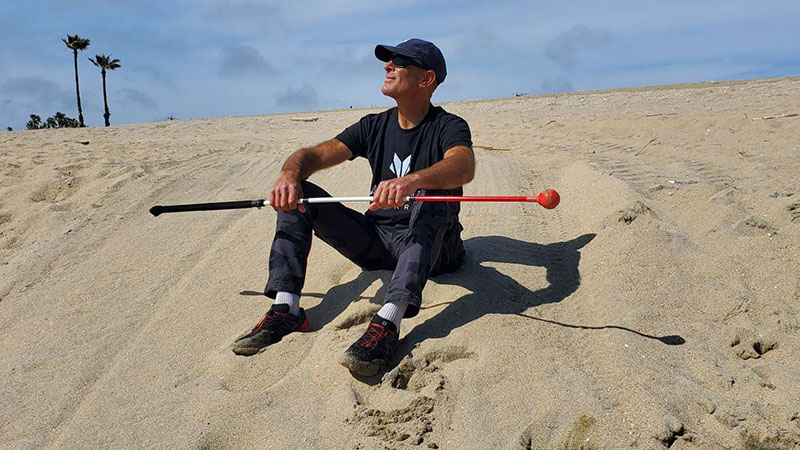 A man sitting outdoors, on a sand dune, wearing sneakers and a baseball cap while holding the Awarewolf outdoor cane.