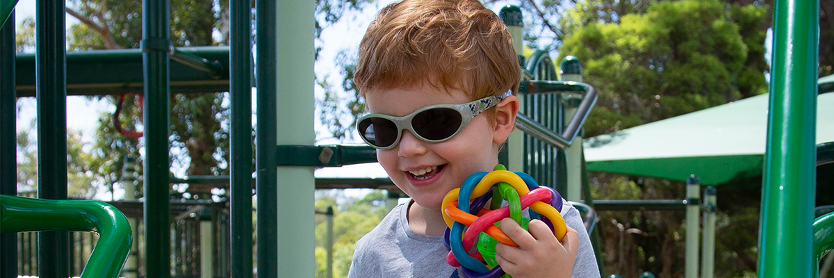 Hugo is smiling at the top of the play equipment. He is holiding a colourful tactile ball and wearing sunglasses
