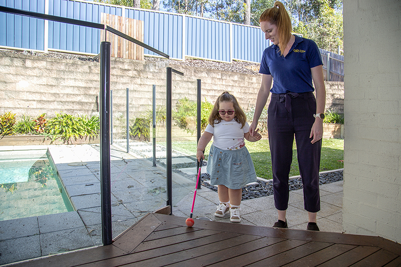 Young client Daisy walks while holding the hand of a Vision Australia staff member and using a cane