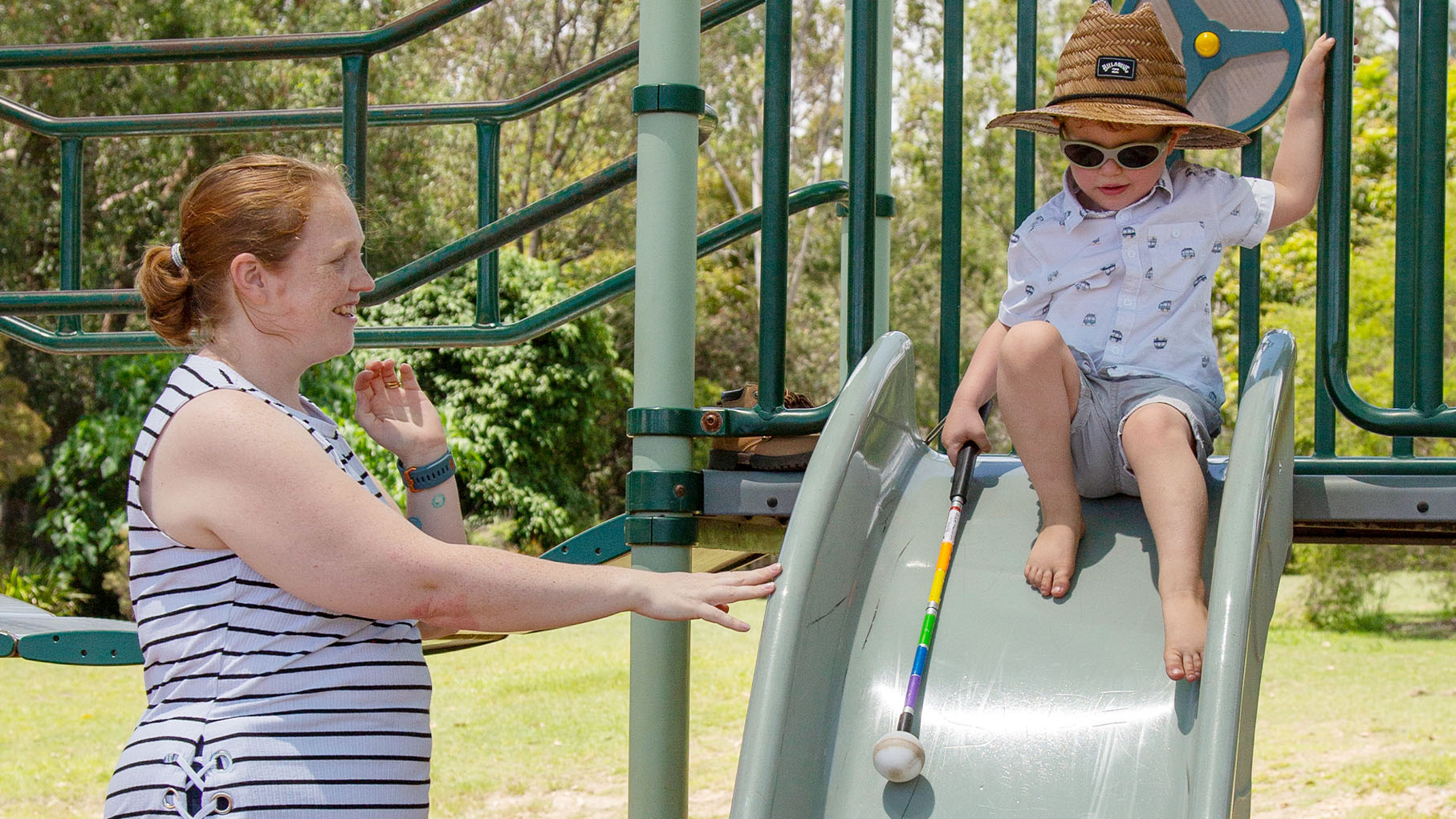 Hugo prepares to slide down the slide, holding his white cane, and his mum is by his side