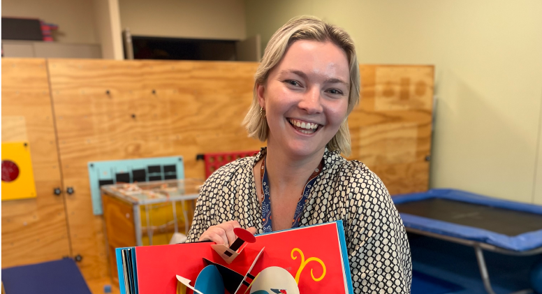 Jemma, Paediatric Speech Pathologist A young woman with short blonde hair smiles at the camera while holding open a pop-up book