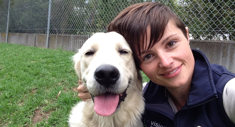 Kat, Senior Kennel Attendant/ Veterinary Nurse A young woman with short brown hair is smiling at the camera while crouched on the ground and resting her head against a yellow dog