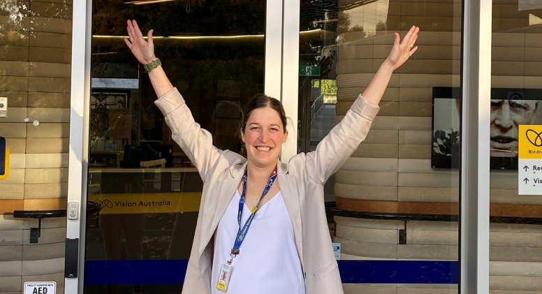 Melissa, People and Culture Business Partner A young woman with tied-up brown hair stands in front of glass doors at Vision Australia Kooyong, smiling at the camera and holding her hands up in the air
