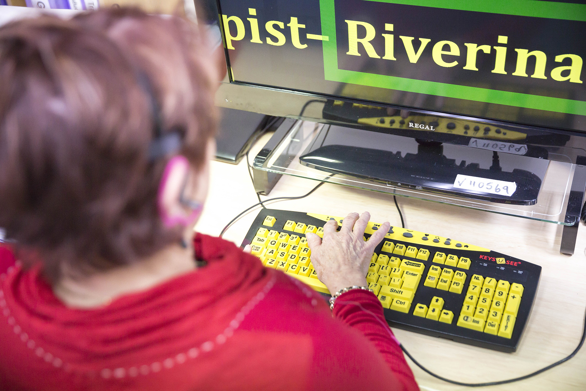 A woman with short hair uses a keyboard with bright yellow letters to type on a computer. On the screen, the word 'Riverina' is shown in large, bright yellow letters and is inside a bright green border