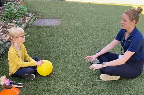 Amelia and Vision Australia occupational therapist seated on the ground and rolling a yellow ball to each other