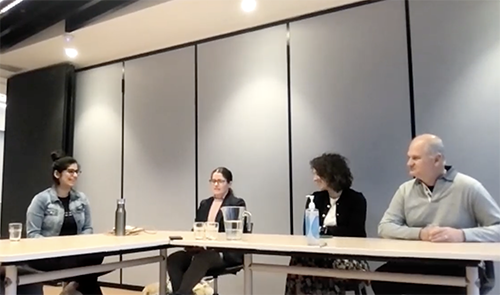 4 people sitting at an L shaped table against a grey wall, 3 of the people have their head turned towards the person in the middle while they listen to them speaking and are smiling.