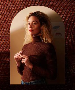 A woman stands side on with her eyes focused towards the left. She has wavy, shoulder length, blonde hair and wears a brown polo neck jumper tucked into blue jeans.