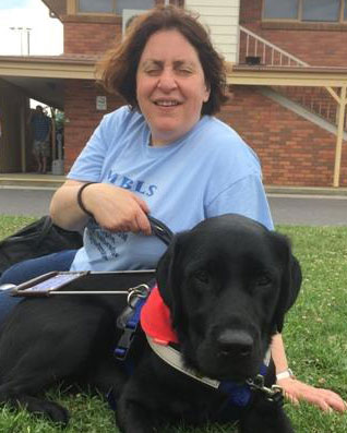 Nadia is sitting on the grass with her black Labrador Seeing Eye Dog, Olympia, sitting in front of her.
