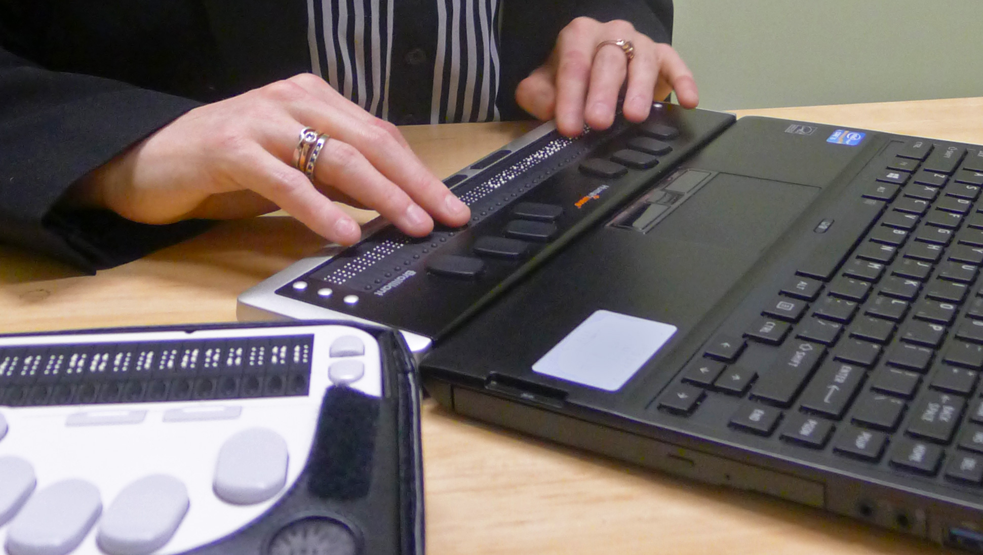 A person's hands reading a refreshable braille display, and a braille note taker device is also in scene.