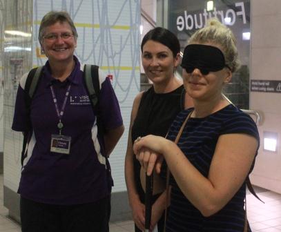 A member from the department of Transport and Main roads wearing a blindfold and holding a cane, standing next to colleague and Vision Australia orientation and mobility specialist.