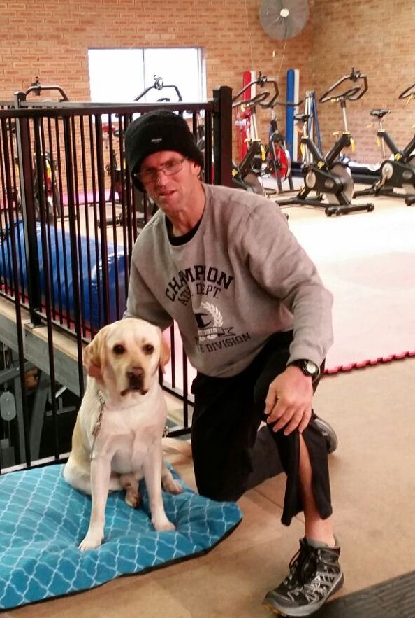 Grant Baker Grant is wearing gym clothes and is leaning down next to his Seeing Eye Dog in shot, with gym equipment in the background