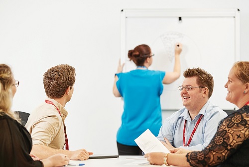 A group of colleagues sit at a table as a woman writes on a whiteboard