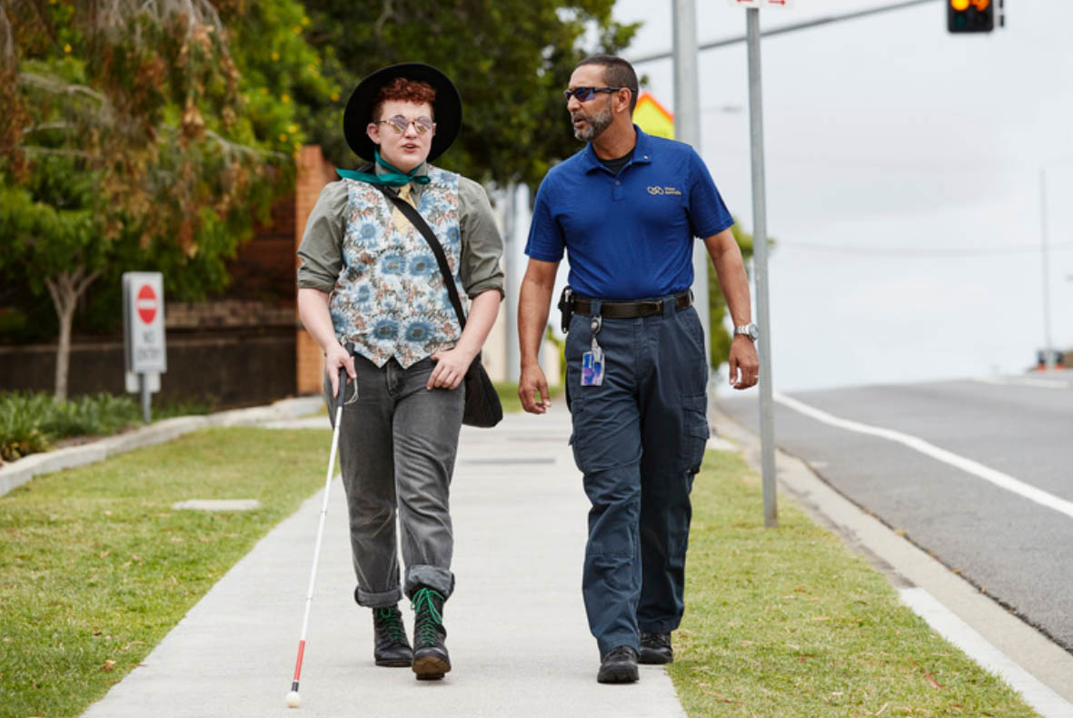 A young man walking with his white cane, walking alongside a staff from Vision Australia.