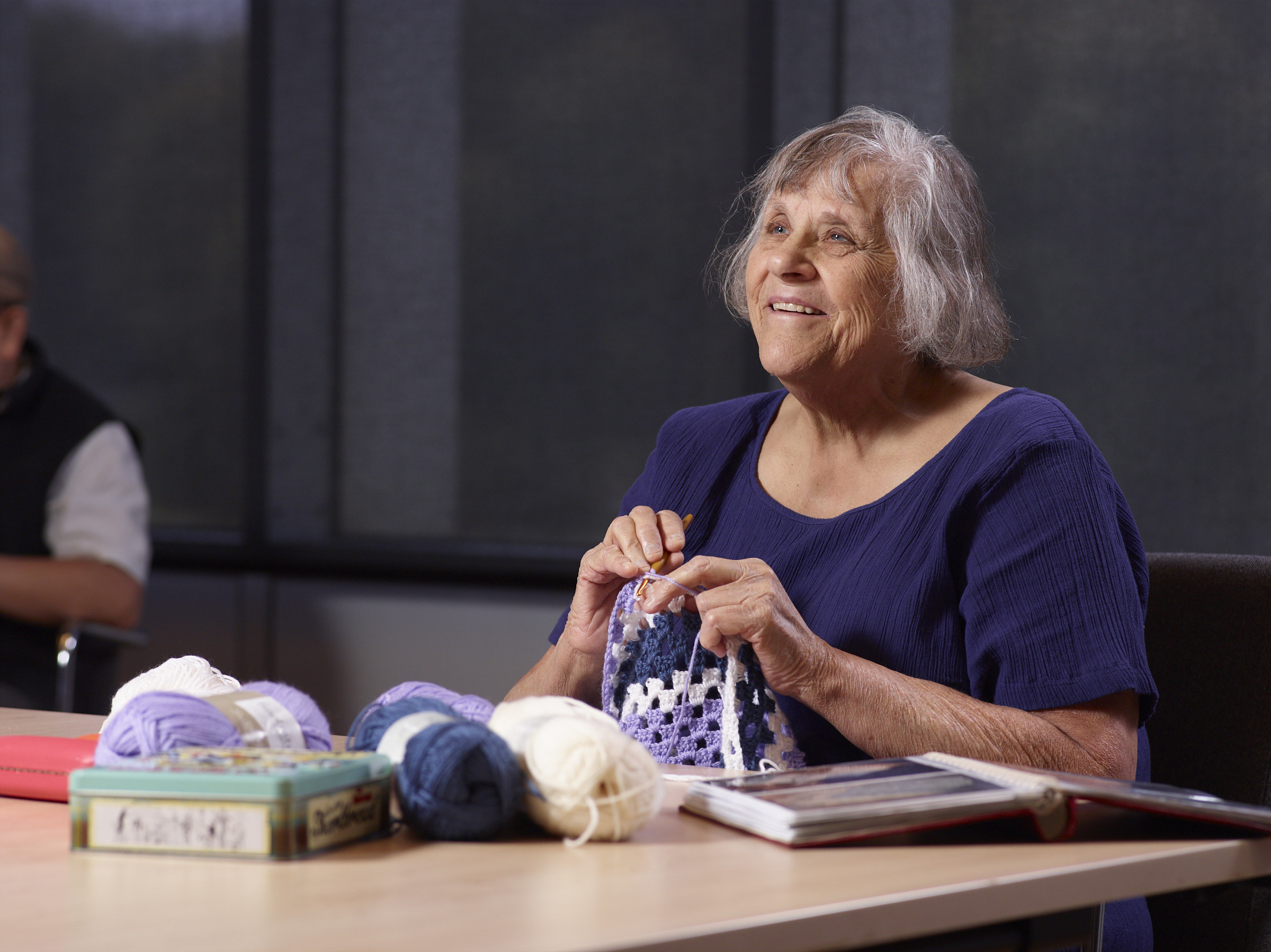 Sarah, an older adult with short grey hair, sits at a table surrounded by thread as she knits