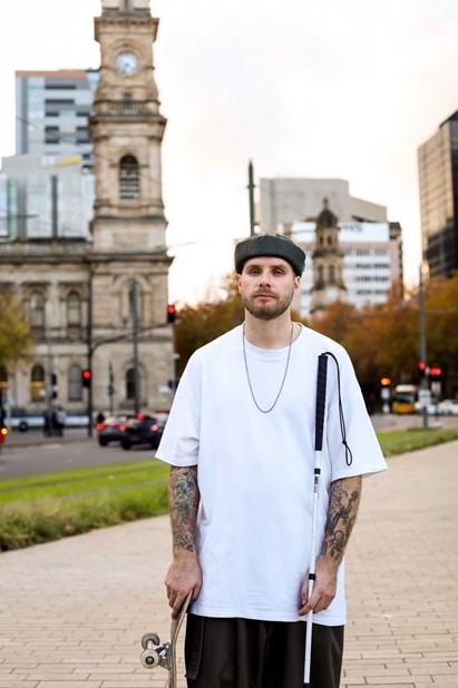 Richard with his cane and skateboard. He is wearing a white top and a grey hat. He has tattoos on both of his arms.