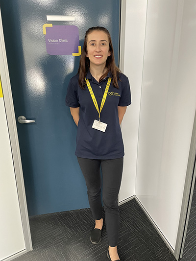 Sekinda has long brown hair and is wearing a navy Vision Australia polo t-shirt. She is smiling and standing in front of her clinic door.