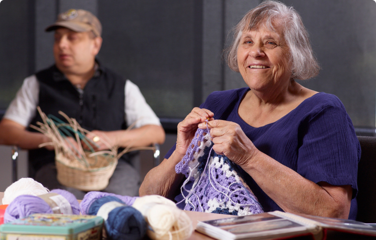 Woman smiling in foreground knits a blkanket, man in background weaves a basket