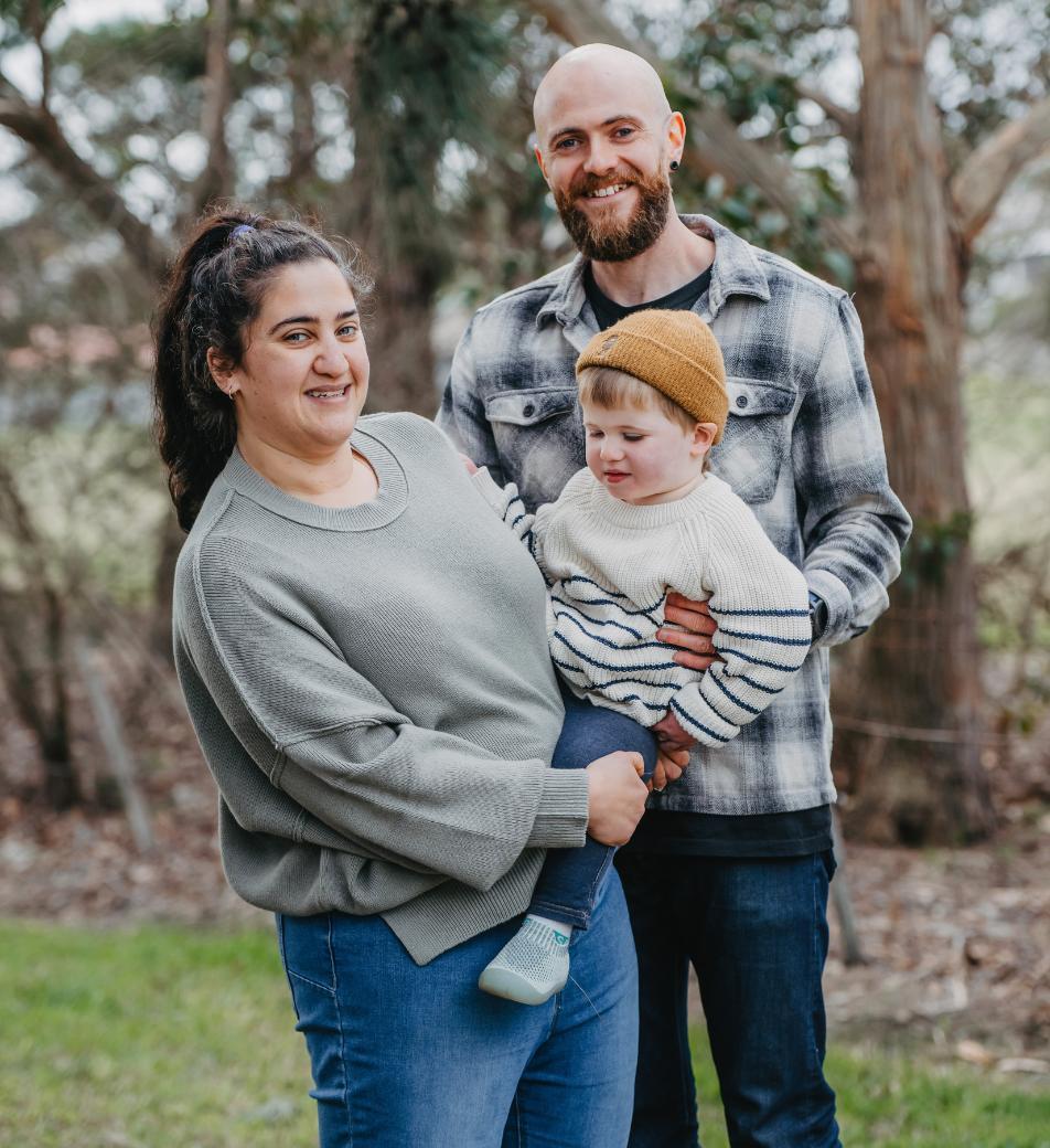 A mum, dad and young child are standing outside, trees are in the background, smiling to camera.