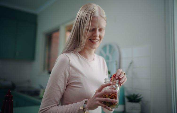 A blonde woman wearing a pink top stands in her kitchen, holding a jar filled with rubber bands.