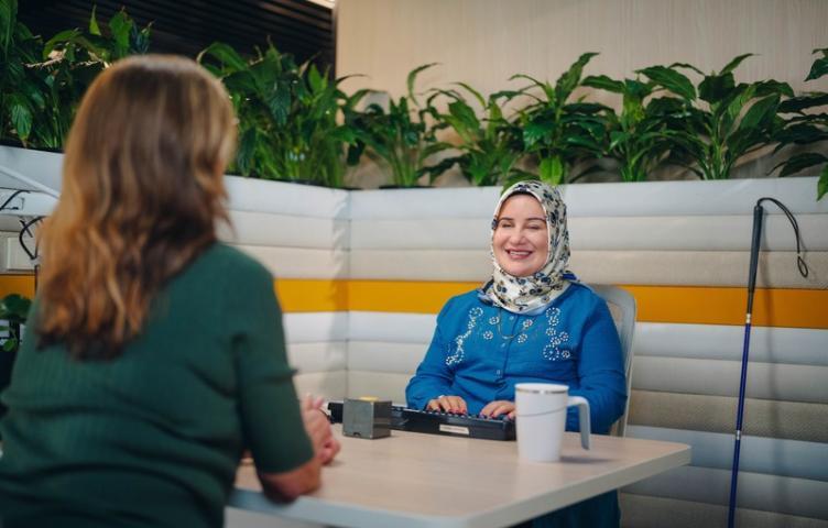 A woman sits at a table typing, her service provider seated across from her, cane resting beside her.