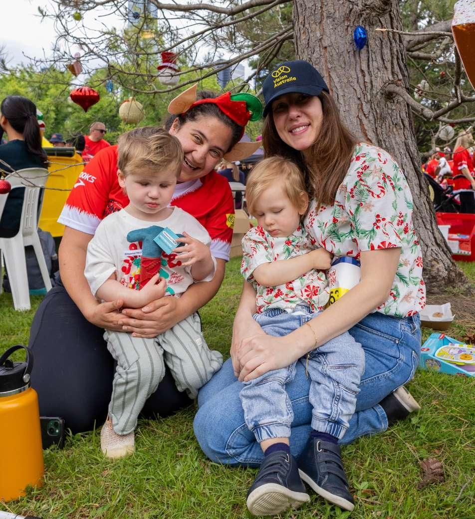 Two women wearing christmas shirts are kneeling on the ground, holding two small children.