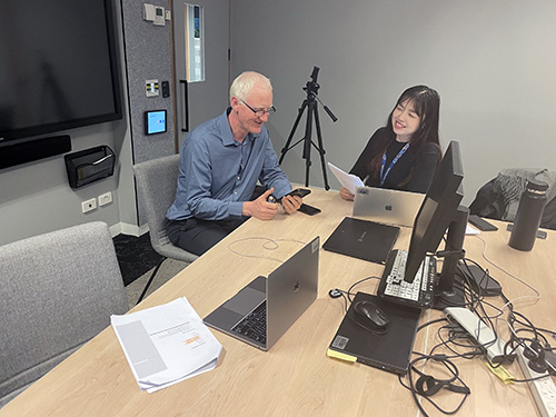 Low vision participant Grant Is looking at his mobile phone and smiling while sitting at a desk next to the Digital Access User Experience consultant Hee-Won who is holding paper with notes and smiling at Grant during a usability testing session. Grant has grey short hair, glasses, light skin, blue business shirt and grey pants. Hee-Won has black long hair, light skin and black long sleeve top wearing a lanyard. There are lots of electronic equipment in front of them on the desk including different computers and cables