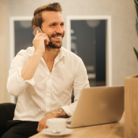 A man smiles while on the phone. He is sitting at a table with a laptop and a coffee cup in front of him.